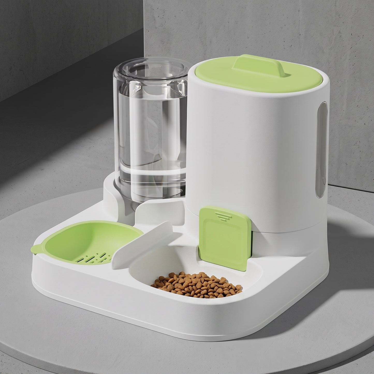 A white automatic pet feeder and water dispenser with light green accents, shown on a round gray table against a gray wall. The feeder includes a clear water bottle, a food hopper filled with kibble, and a green bowl.