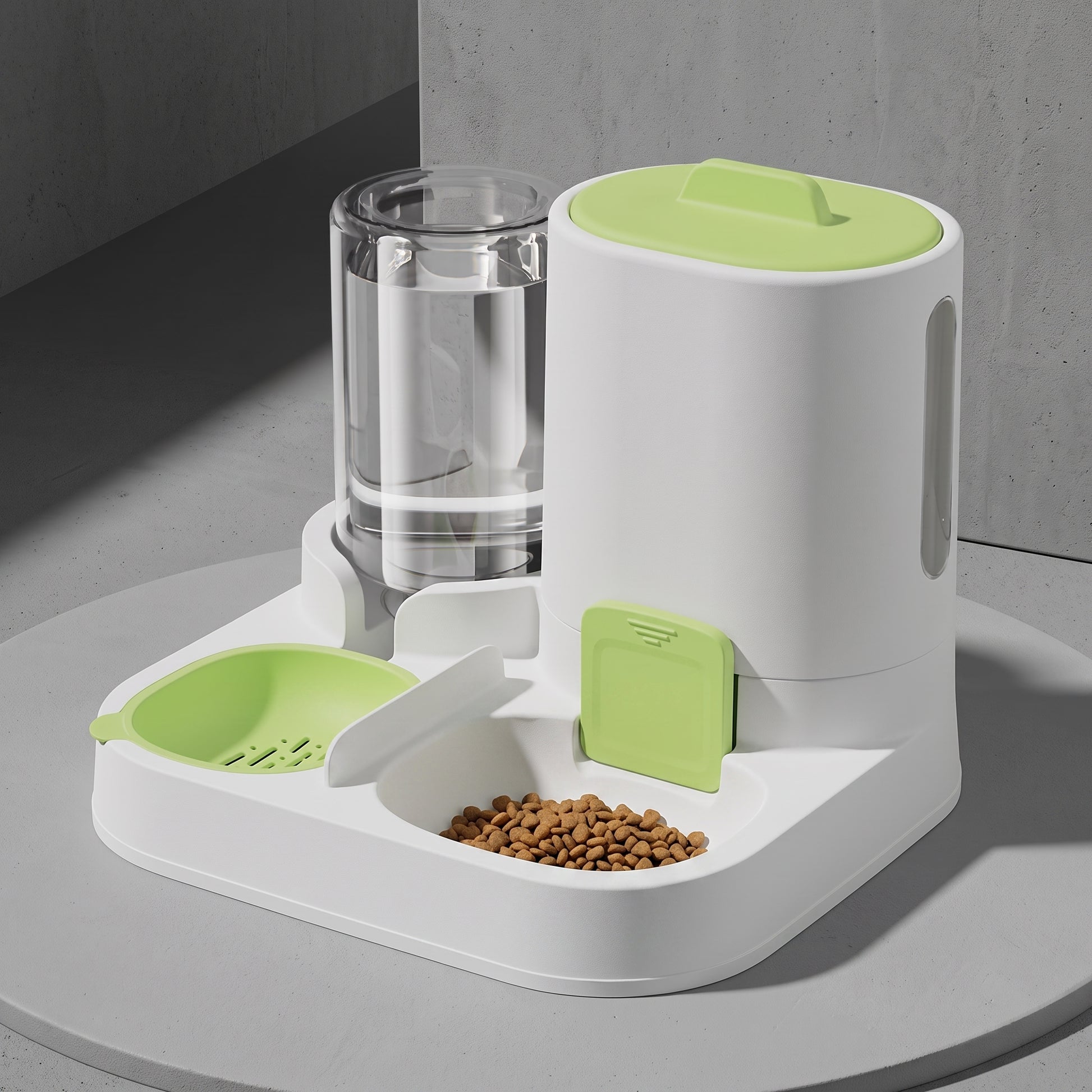 A white automatic pet feeder and water dispenser with light green accents, shown on a round gray table against a gray wall. The feeder includes a clear water bottle, a food hopper filled with kibble, and a green bowl.