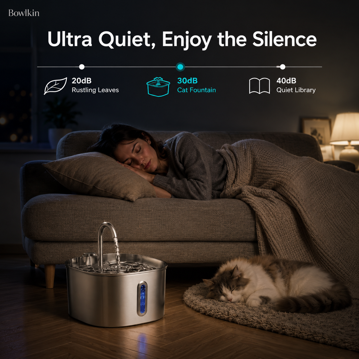 A cat fountain by Bowkin shown in a cozy living room scene with a sleeping woman on a couch and a cat on the floor. The product is a stainless steel cat fountain with a water reservoir and faucet, positioned on the floor near the cat.