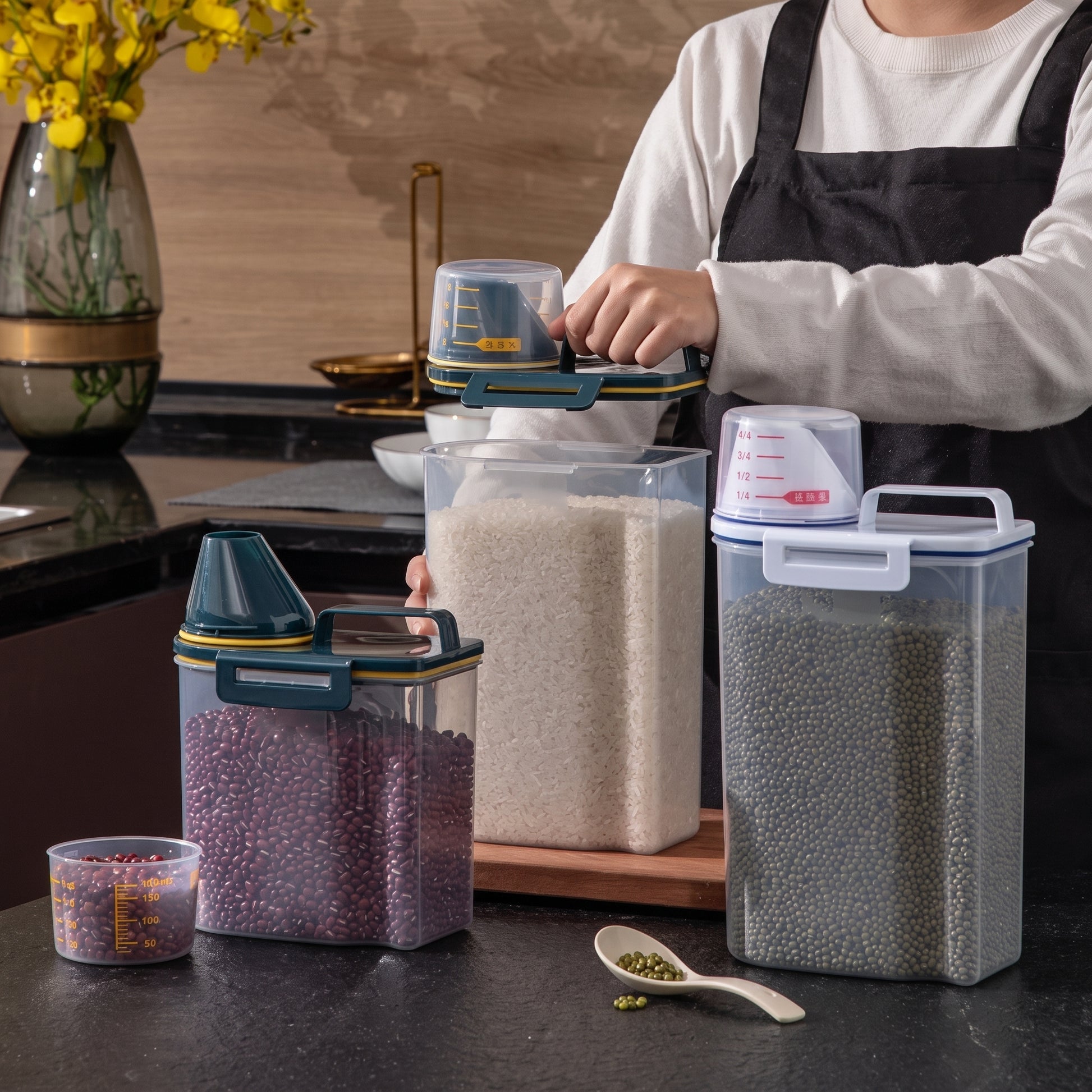 Three clear plastic grain storage containers with lids and measuring cups are shown on a kitchen counter, with a person in a white shirt and black apron pouring grain into the center container. The containers are filled with rice, lentils, and red beans, and a small clear cup and spoon are placed in front.
