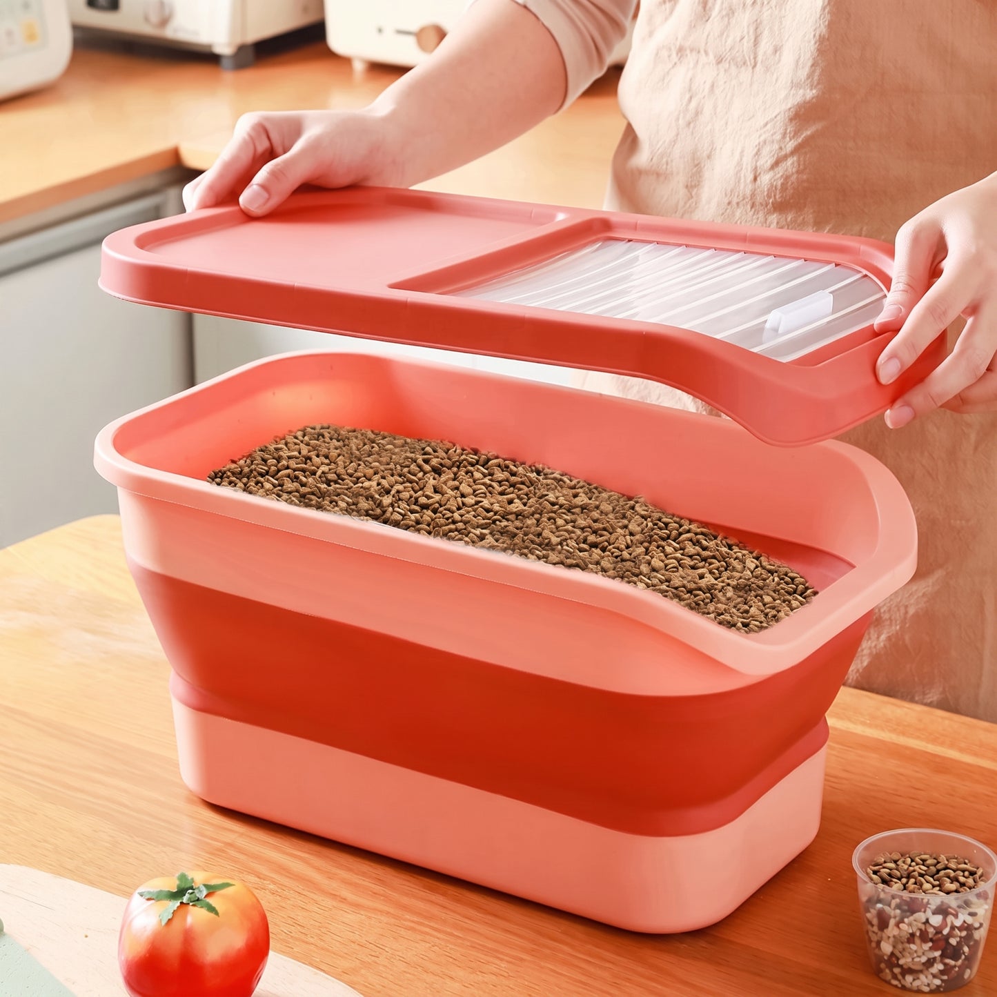 A person in a beige apron is shown holding the lid of a collapsible rectangular container filled with brown granular material, placed on a wooden countertop in a kitchen setting.