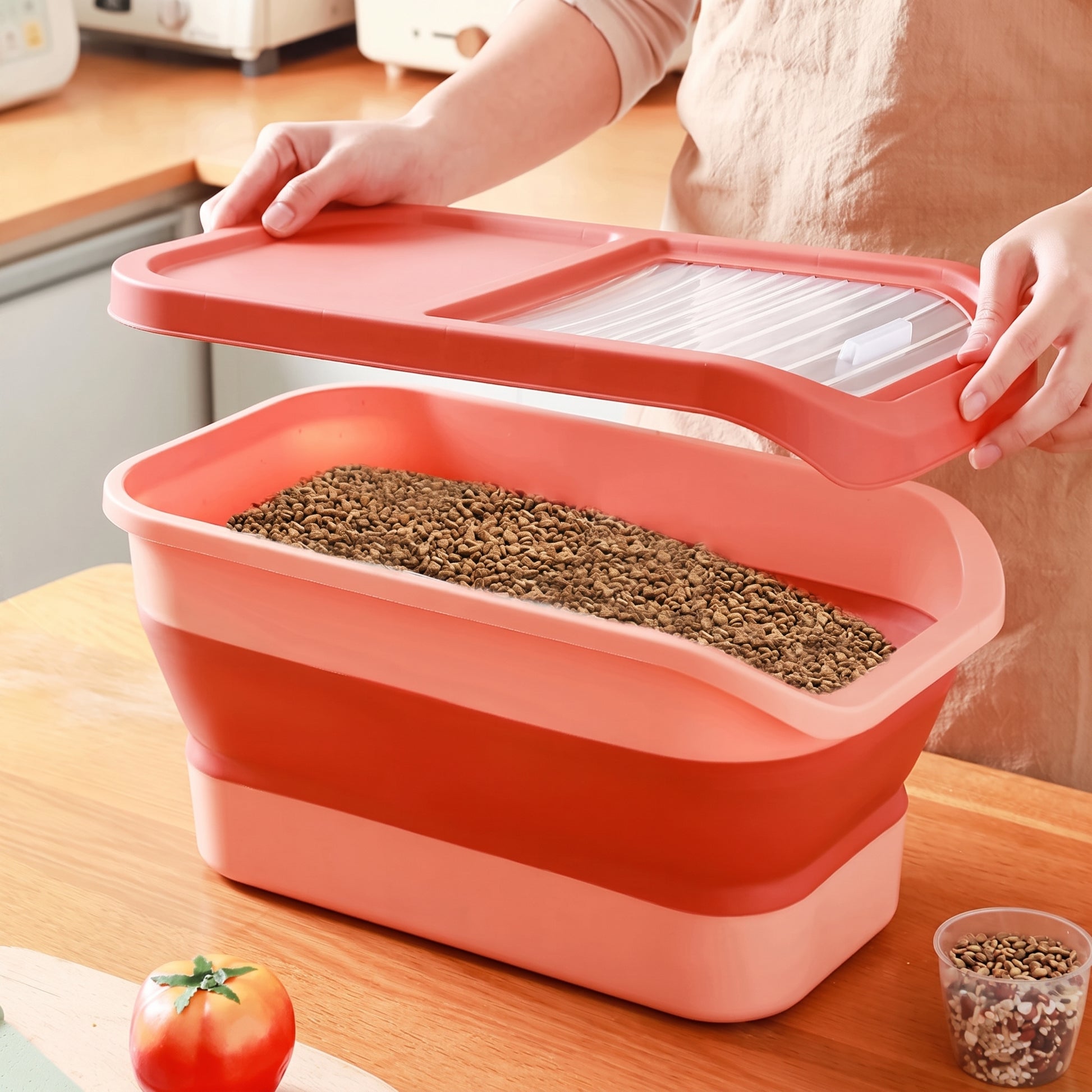 A person in a beige apron is shown holding the lid of a collapsible rectangular container filled with brown granular material, placed on a wooden countertop in a kitchen setting.