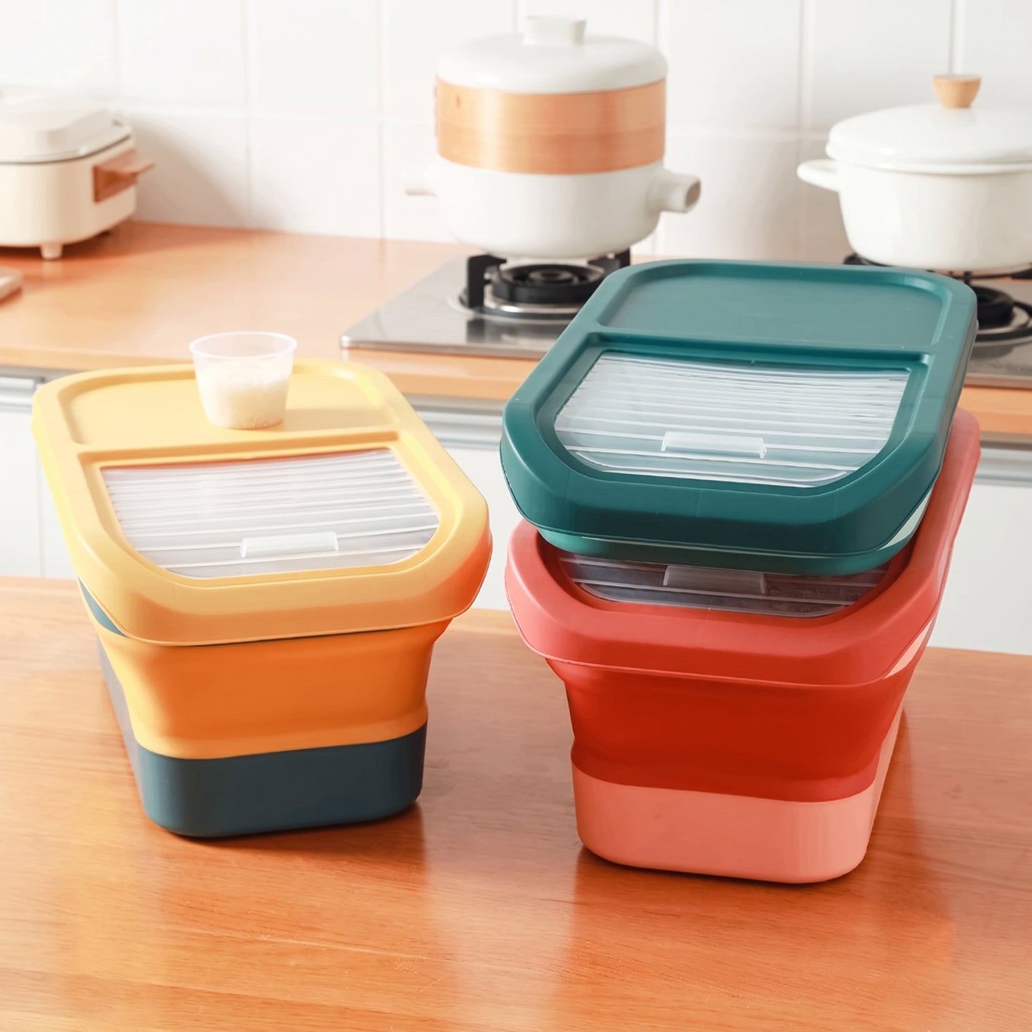 Two stackable collapsible food storage containers with lids are shown on a wooden kitchen counter. One container is yellow and orange with a dark teal base, and the other is coral red and pink with a teal lid.