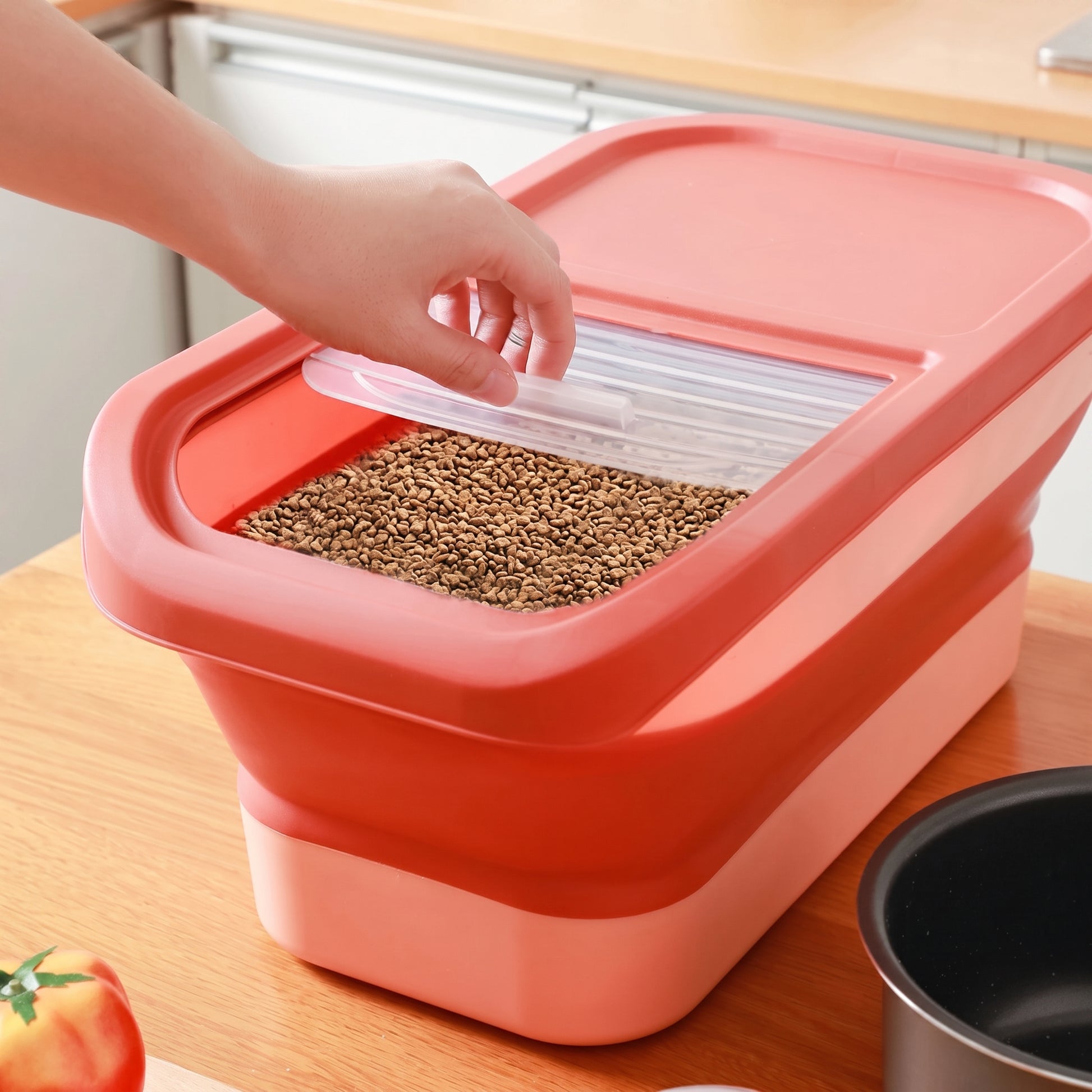 A red and pink collapsible plastic grain bin filled with brown grains, with a hand lifting the clear lid, set on a wooden countertop.