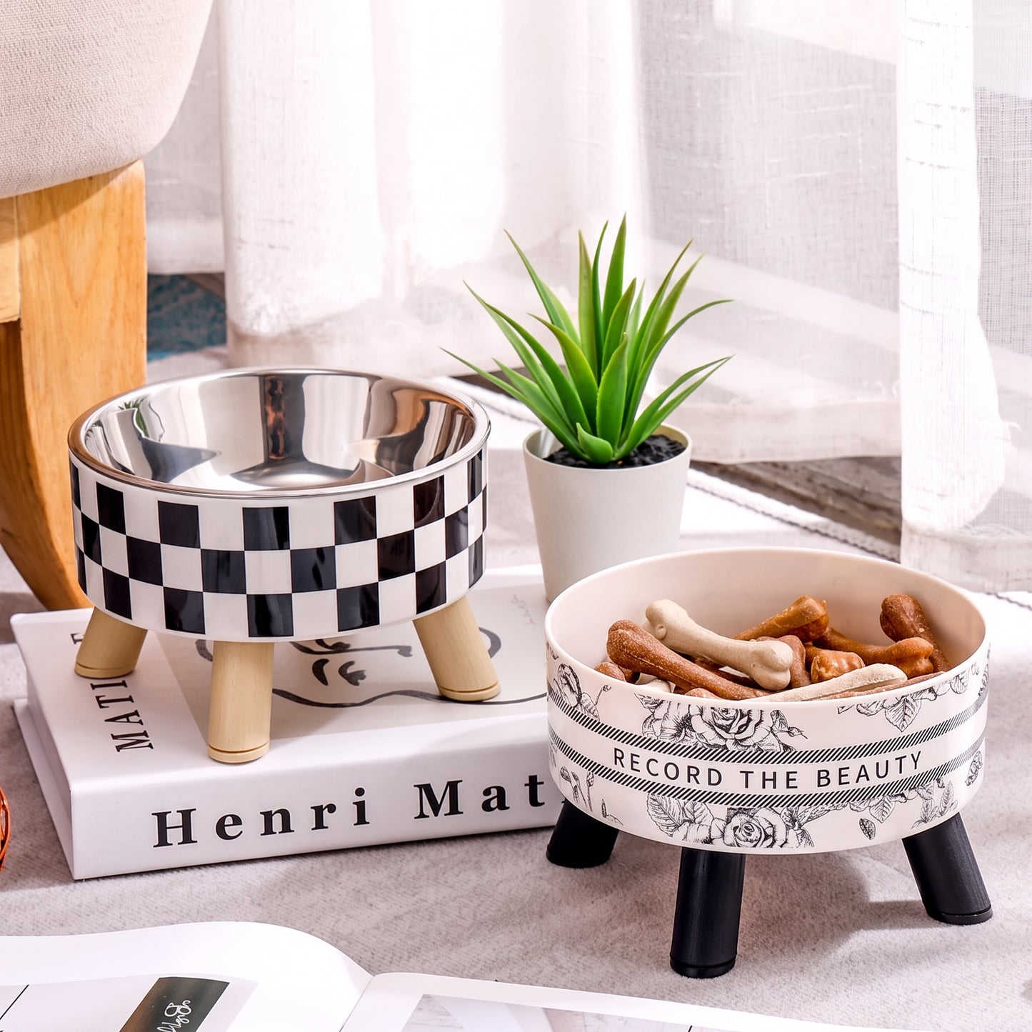 Two decorative elevated pet bowls on a tabletop in a styled indoor setting. One bowl has a black-and-white checkerboard pattern with beige legs, and the other is white with black floral scrollwork and the phrase 'RECORD THE BEAUTY,' containing bones inside.
