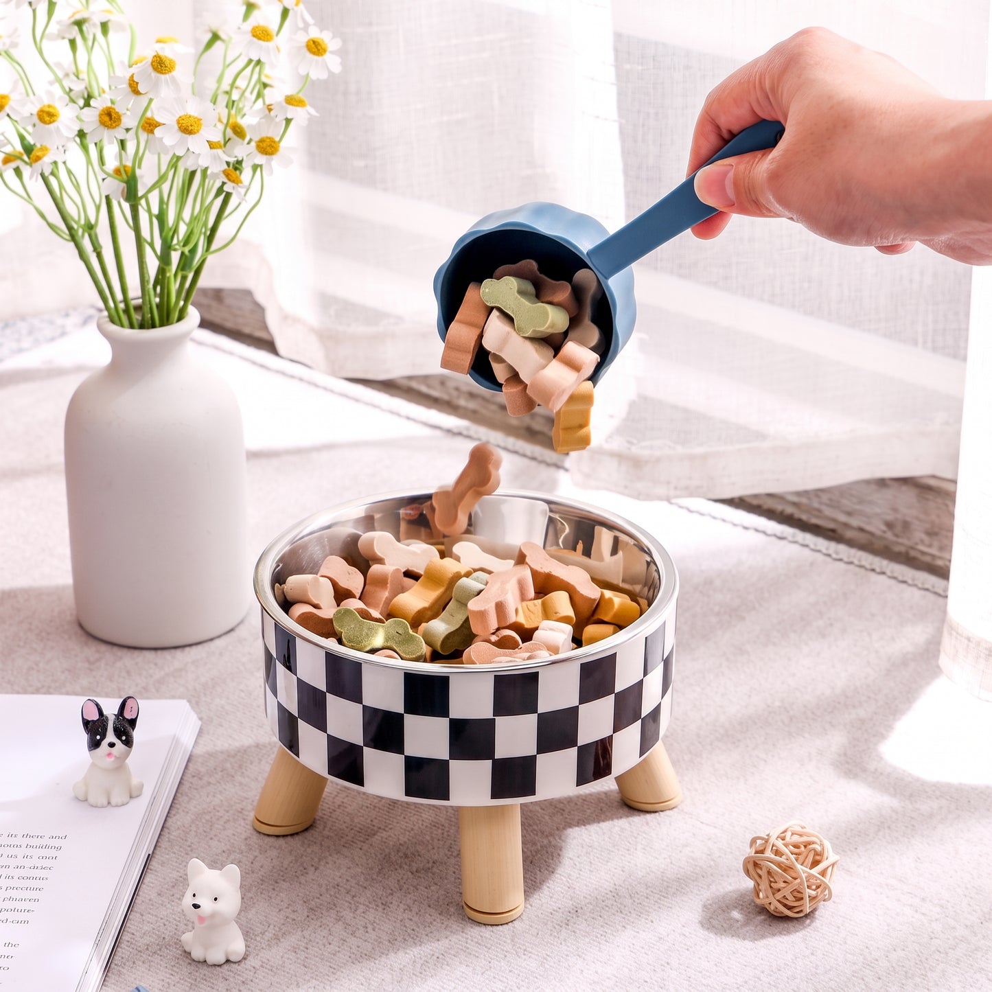 Checkered pet bowl with dog treats being poured from a scoop, next to a vase of flowers and small figurines.