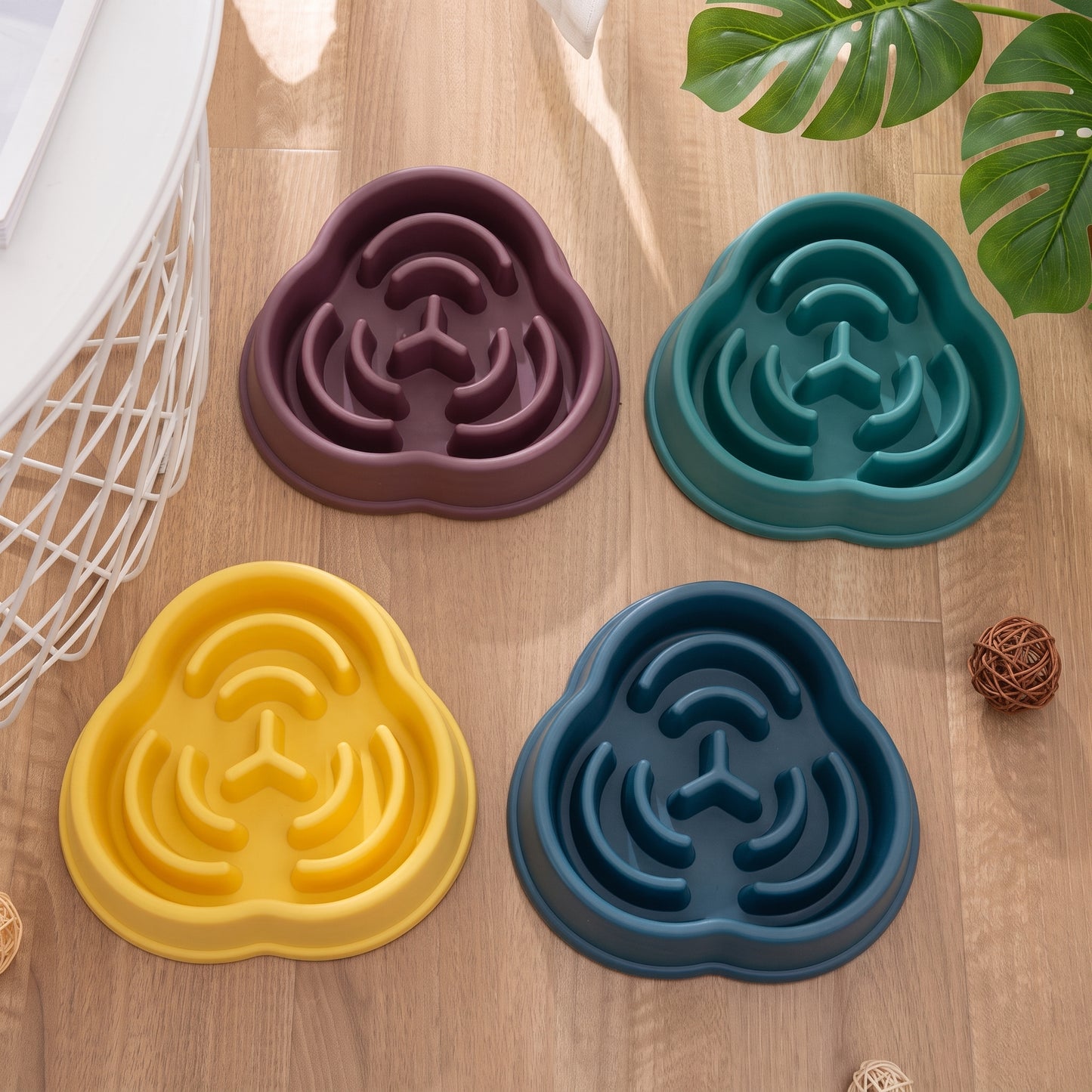 Four cloud-shaped slow feeder dog bowls in purple, teal, yellow, and blue, arranged on a light wood floor with decorative props.