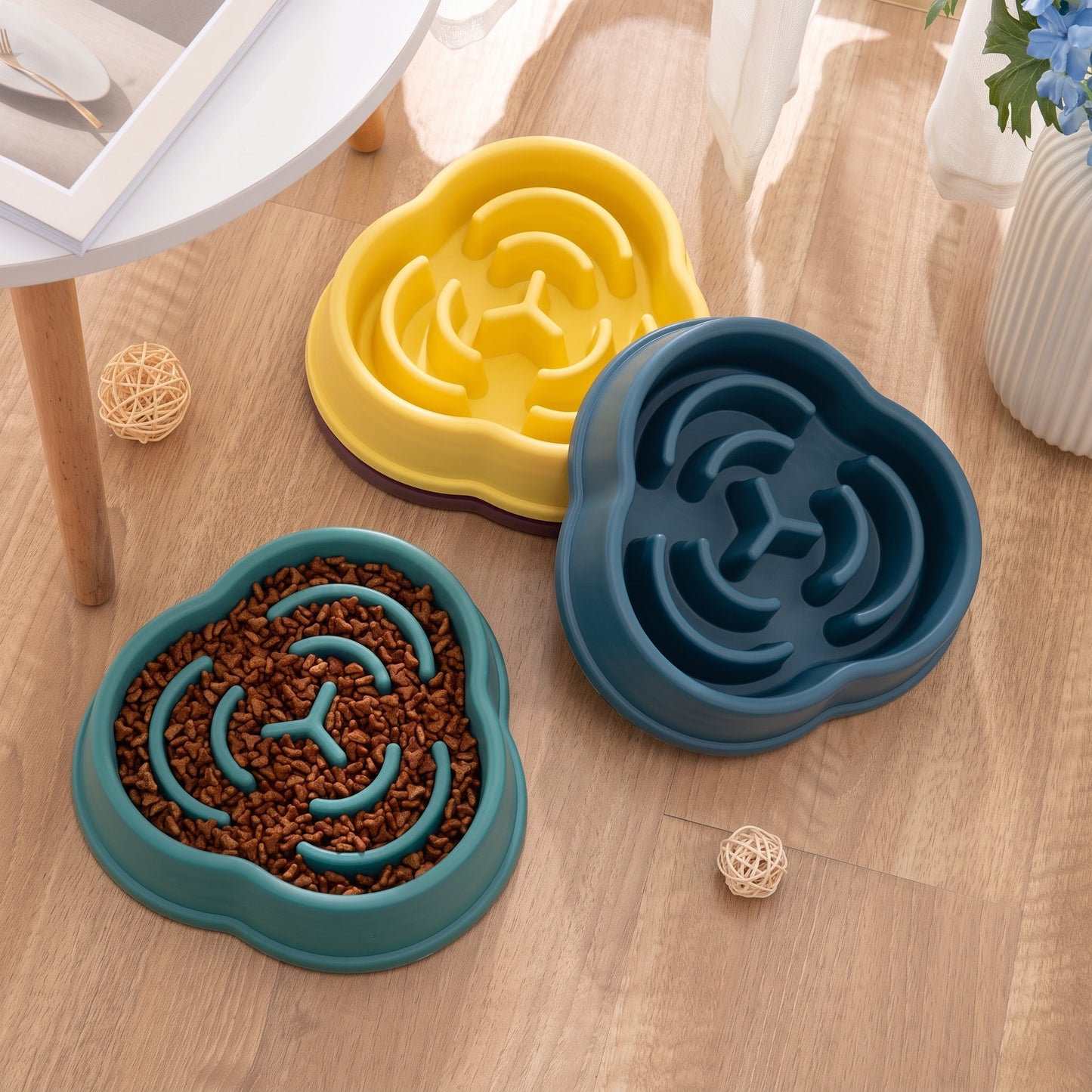 Three slow-feeding pet bowls with a cloud-like shape are arranged on a light wood floor. One bowl is teal and filled with kibble, while the other two are shown empty in yellow and dark teal.