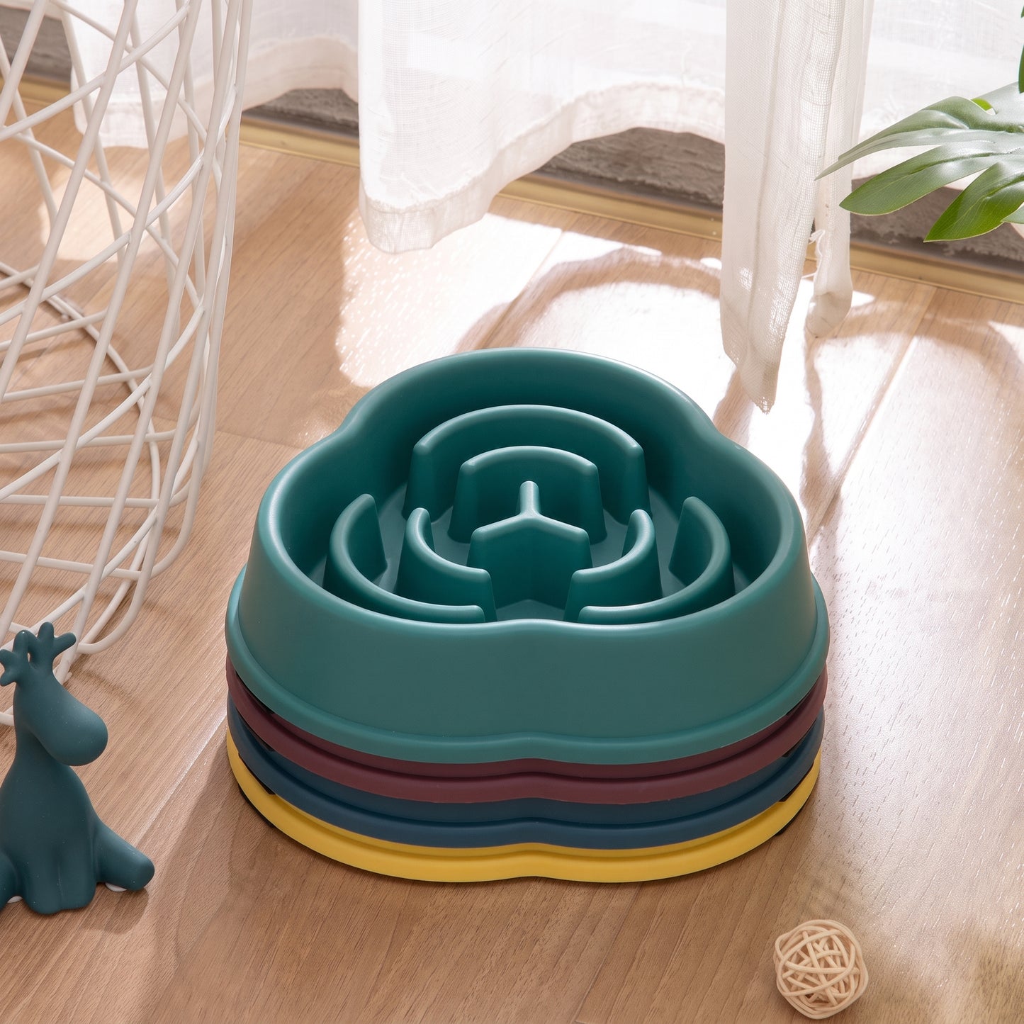 Teal pet slow feeder bowl on a wooden floor with a plant and decorative items in the background